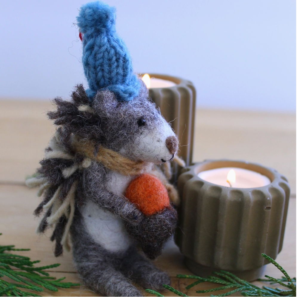 Textile art of a hedgehog with a blue knitted hat and orange scarf, sitting next to a candle on a wooden surface.