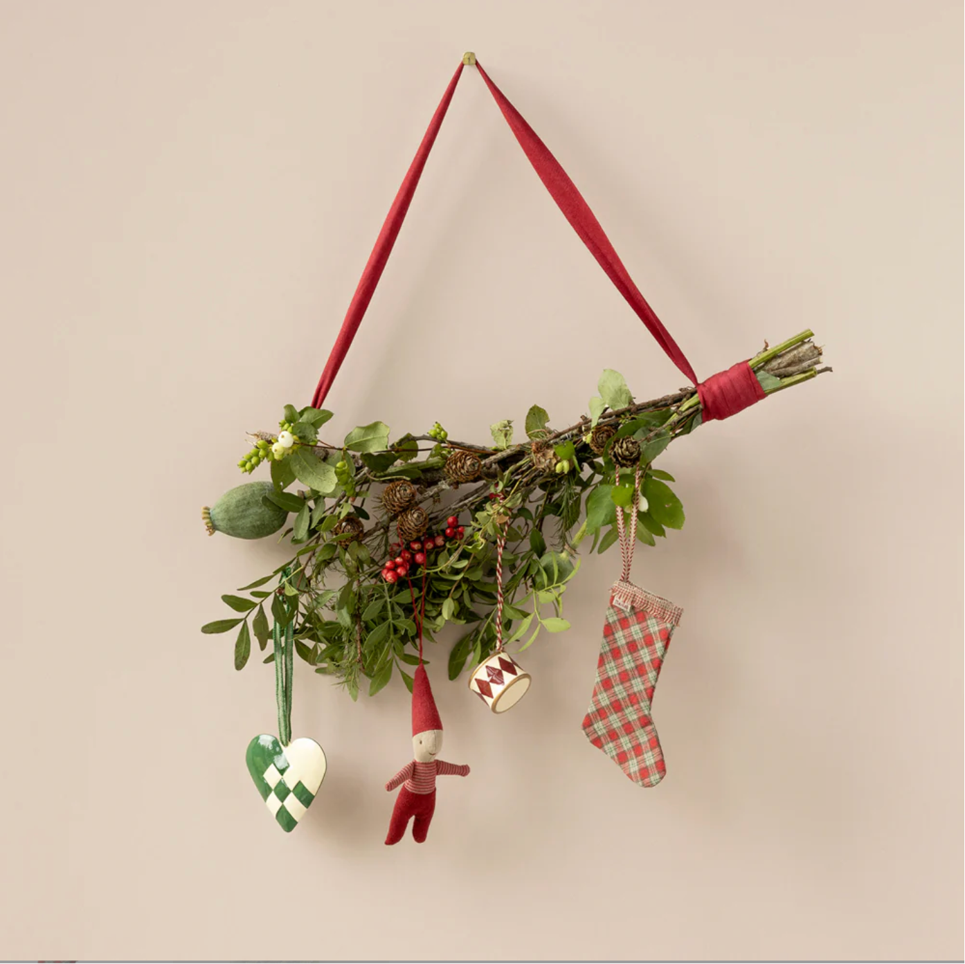 Decorative wall hanging with greenery, small figurines, and red ribbons on a beige background