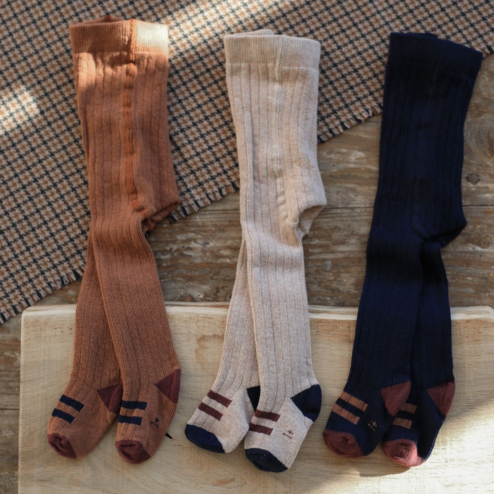 Three pairs of socks in brown, beige, and navy on a wooden surface with a checkered fabric background.