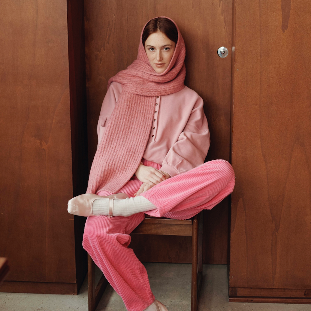 Person wearing a pink outfit sitting on a wooden stool against a wooden wall.
