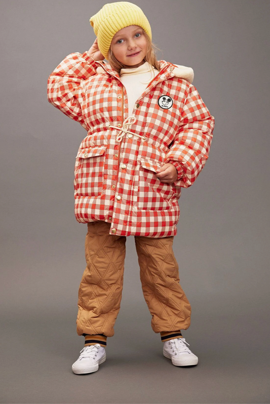 Child wearing a red and white checkered coat with a yellow beanie and brown pants on a gray background