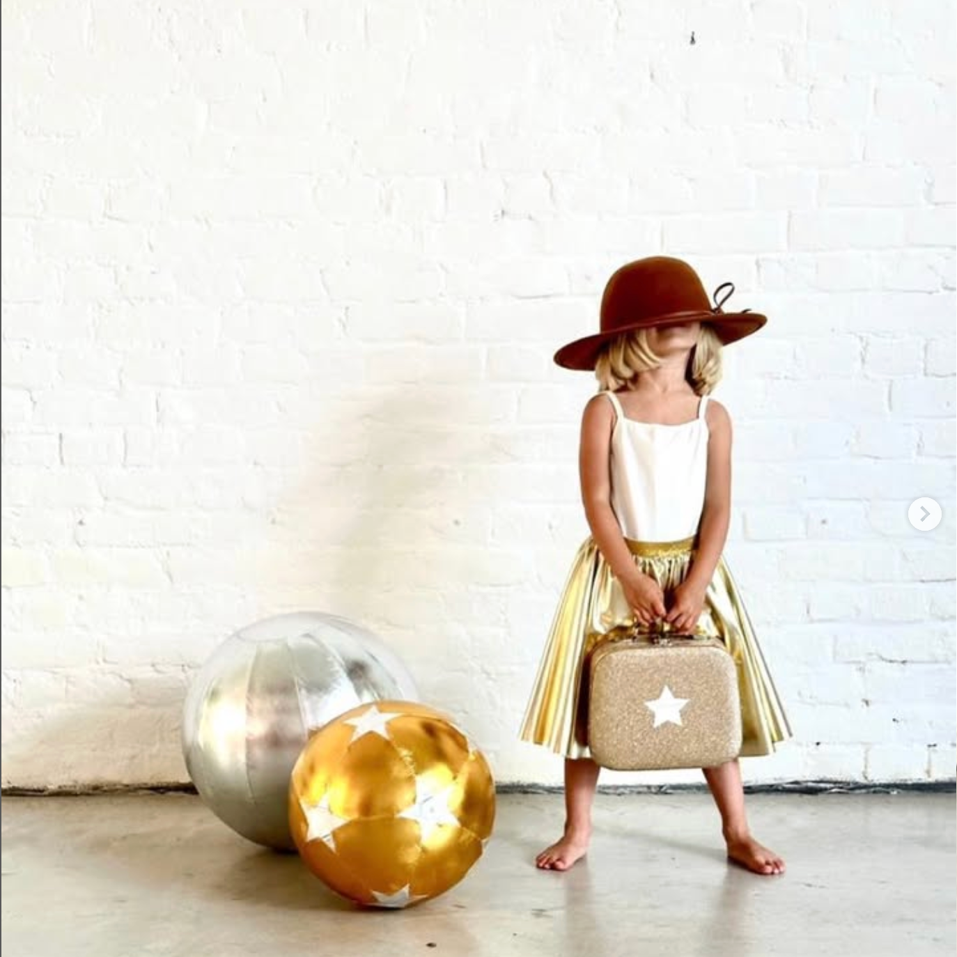 Child in a gold dress and brown hat standing next to decorative balls against a white brick wall.