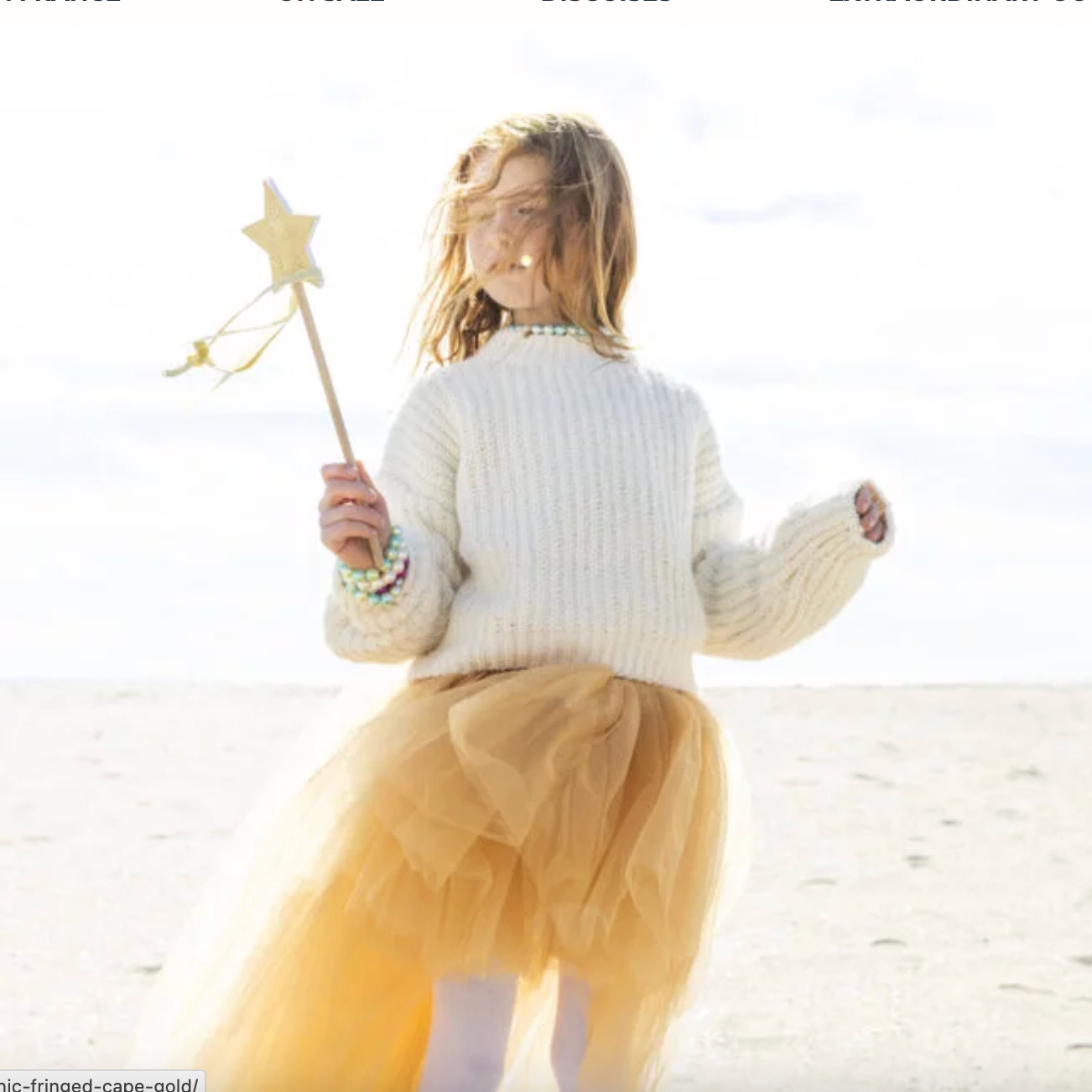 Child in a white sweater and yellow skirt holding a star-shaped wand on a beach.