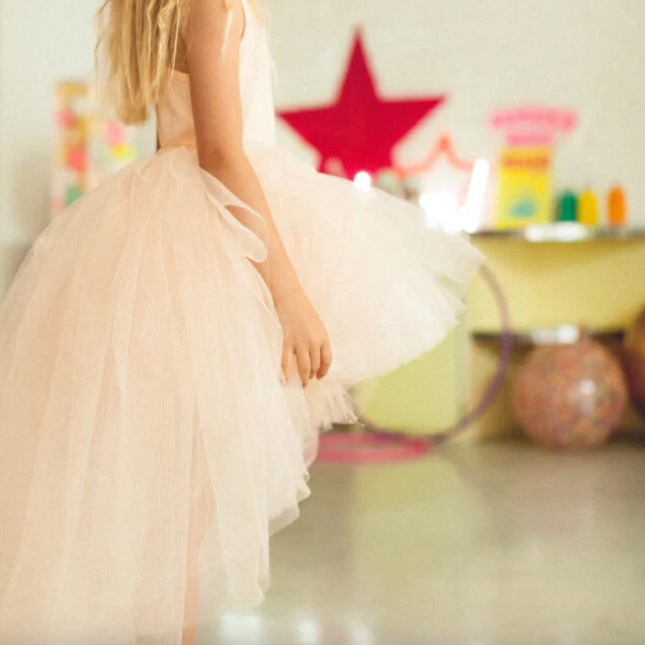 Child in a tutu standing in a room with colorful toys and a red star decoration.