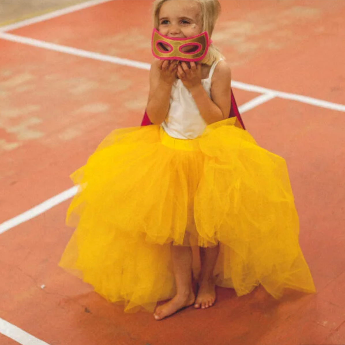 Child in a yellow tutu and pink mask sitting on a track