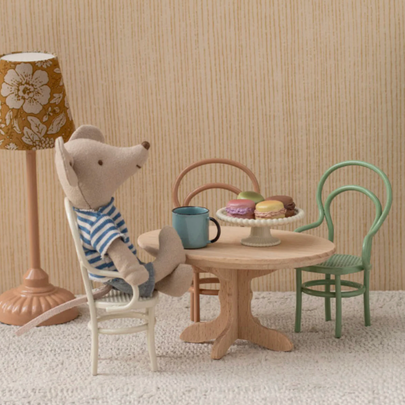 Miniature mouse sitting at a table with a cake and cup, surrounded by chairs and a lamp.